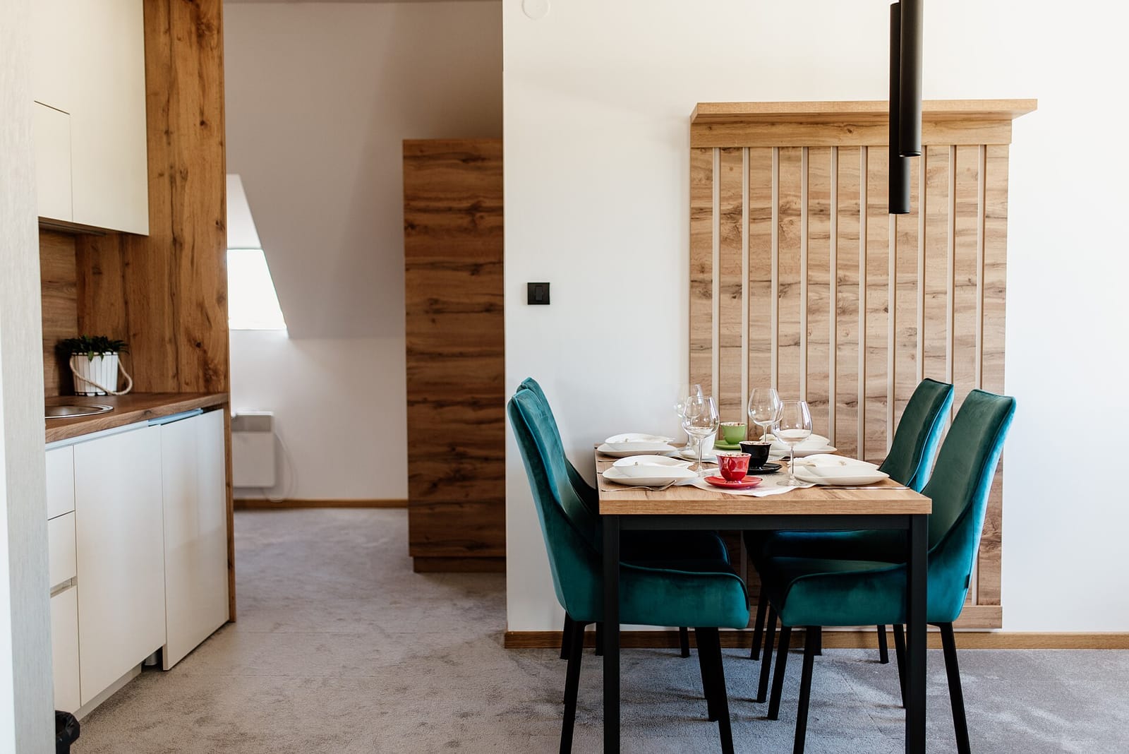 A dining table laid with plates and glasses, surrounded by teal upholstered chairs, with a white kitchen behind it.
