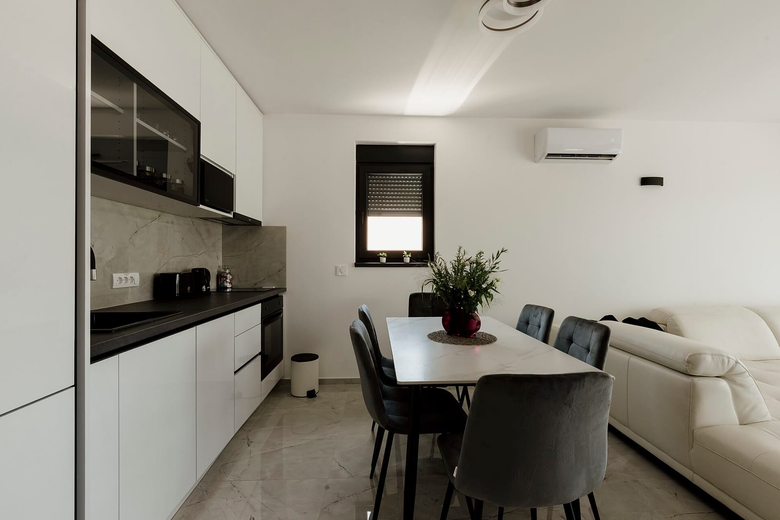 A bright open-plan kitchen and dining area with white gloss units, a dark worktop and a clear glass dining table.