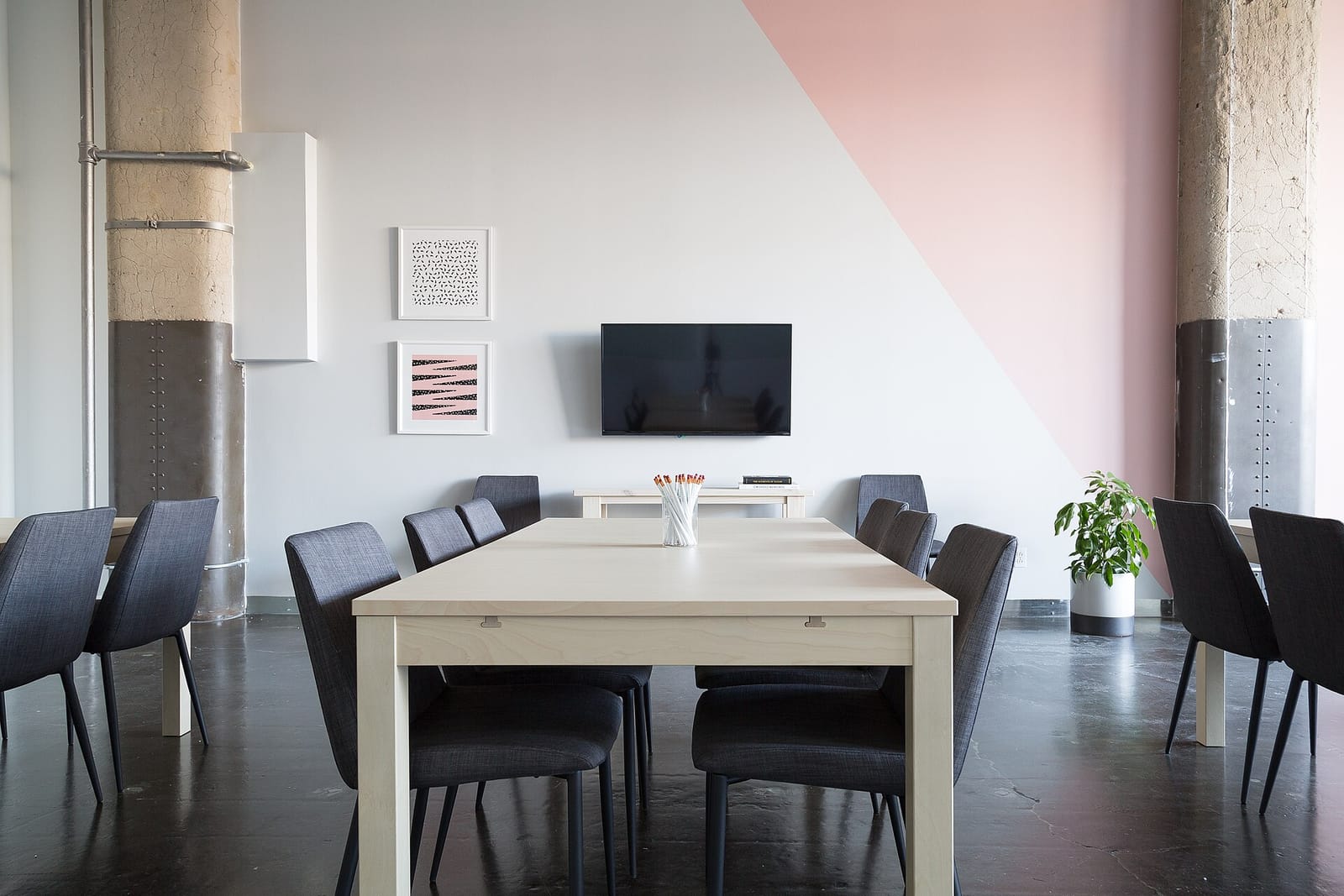 An empty meeting room with a long pale wood table, dark upholstered chairs and a wall-mounted screen.