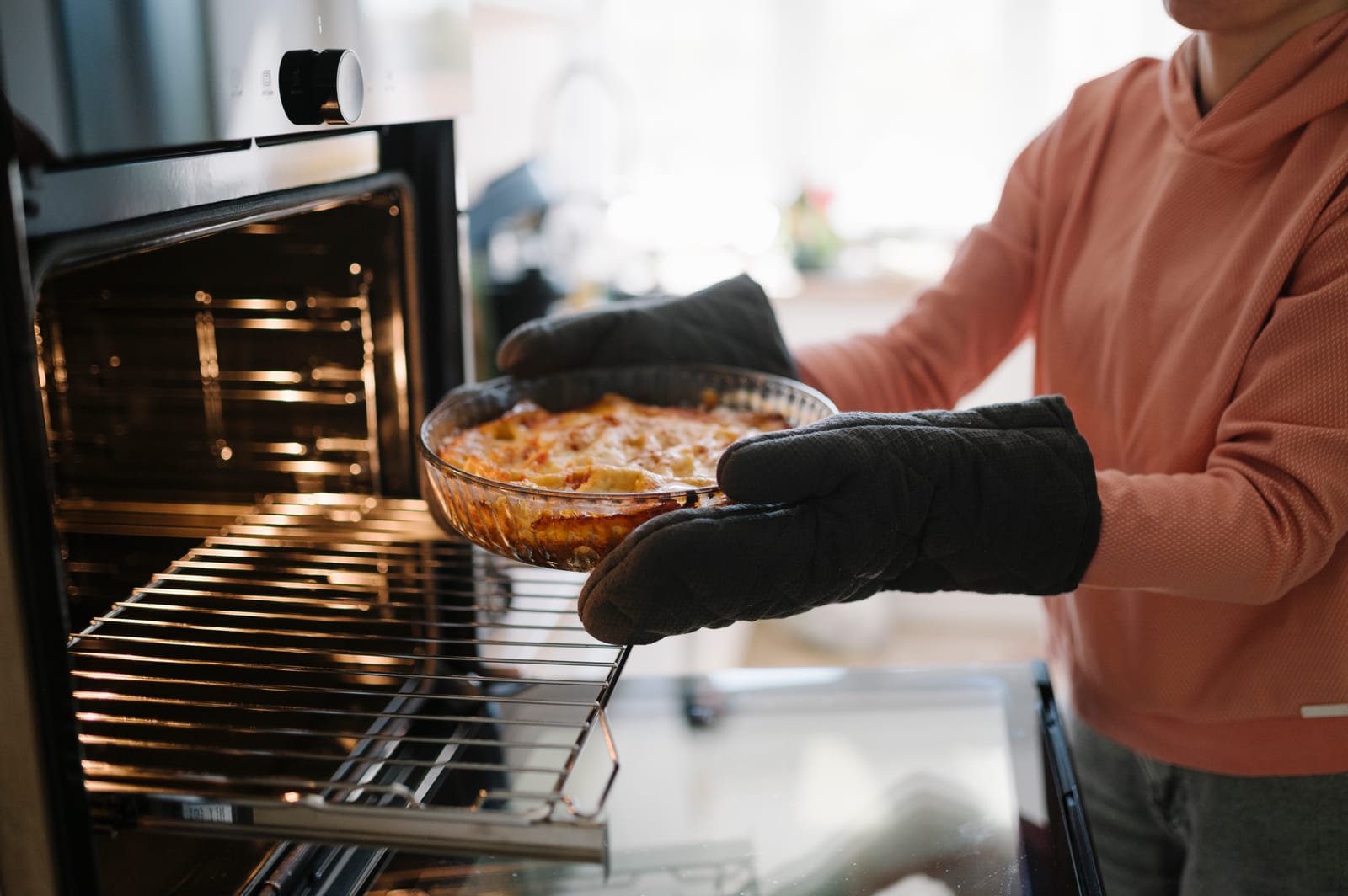 A built-in oven standing open with a clean, gleaming interior and bare shelves, as a baked dish is lifted out on oven gloves.