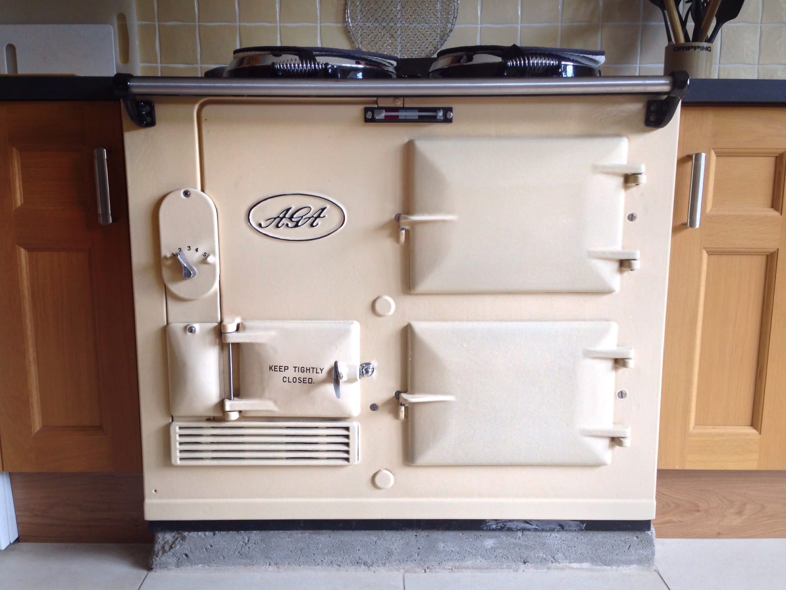 A cream range cooker set between oak kitchen units, its enamel doors, chrome rail and hotplate lids wiped clean.